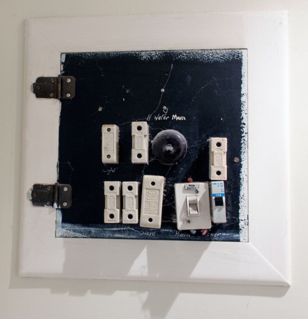 An old domestic electrical switchboard with ceramic fuses, outdated toggle switches, and cobwebs, mounted in a white wooden frame.